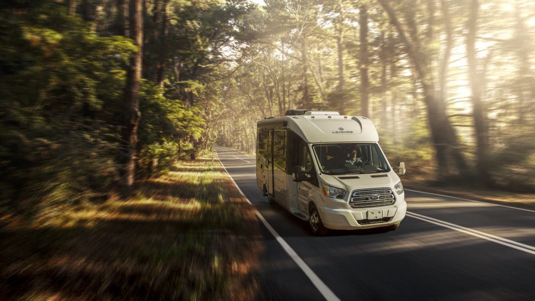 A white motorhome traveling down the road in a forest setting.
