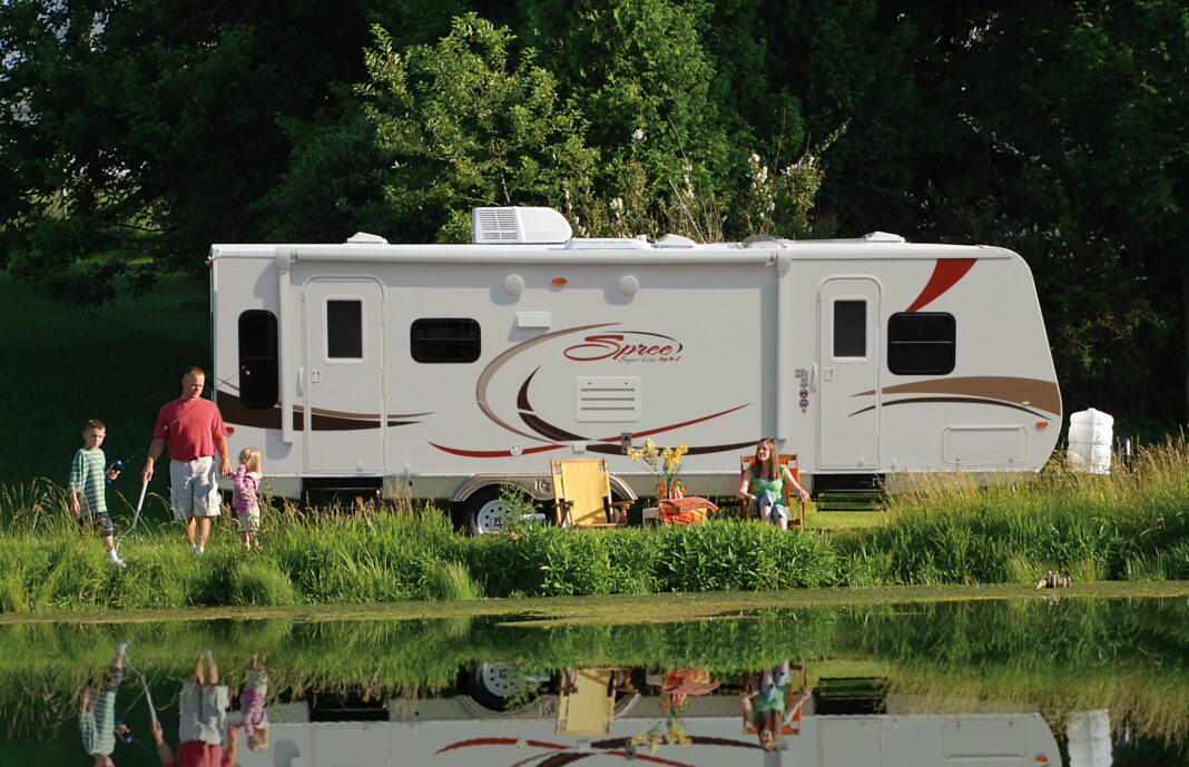 Family enjoying the RV camping experience by a pond at their campground. KZ-RV Spree Ultralight travel trailer, side view.