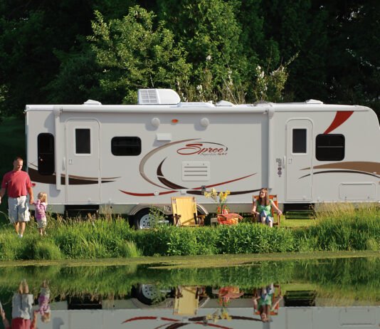Family enjoying the RV camping experience by a pond at their campground. KZ-RV Spree Ultralight travel trailer, side view.