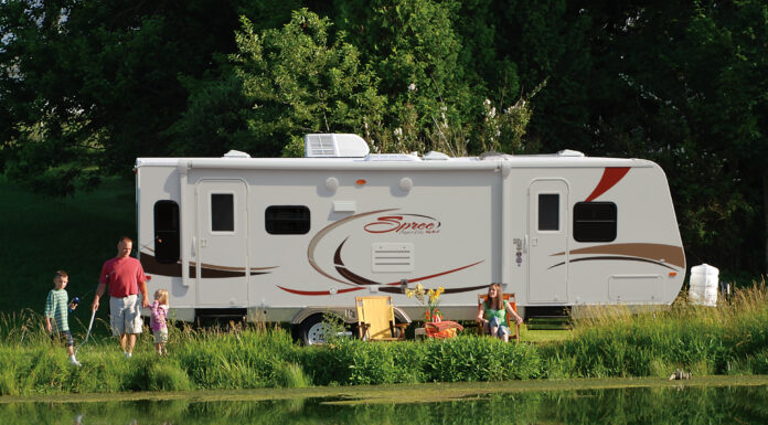 Family enjoying the RV camping experience by a pond at their campground. KZ-RV Spree Ultralight travel trailer, side view.